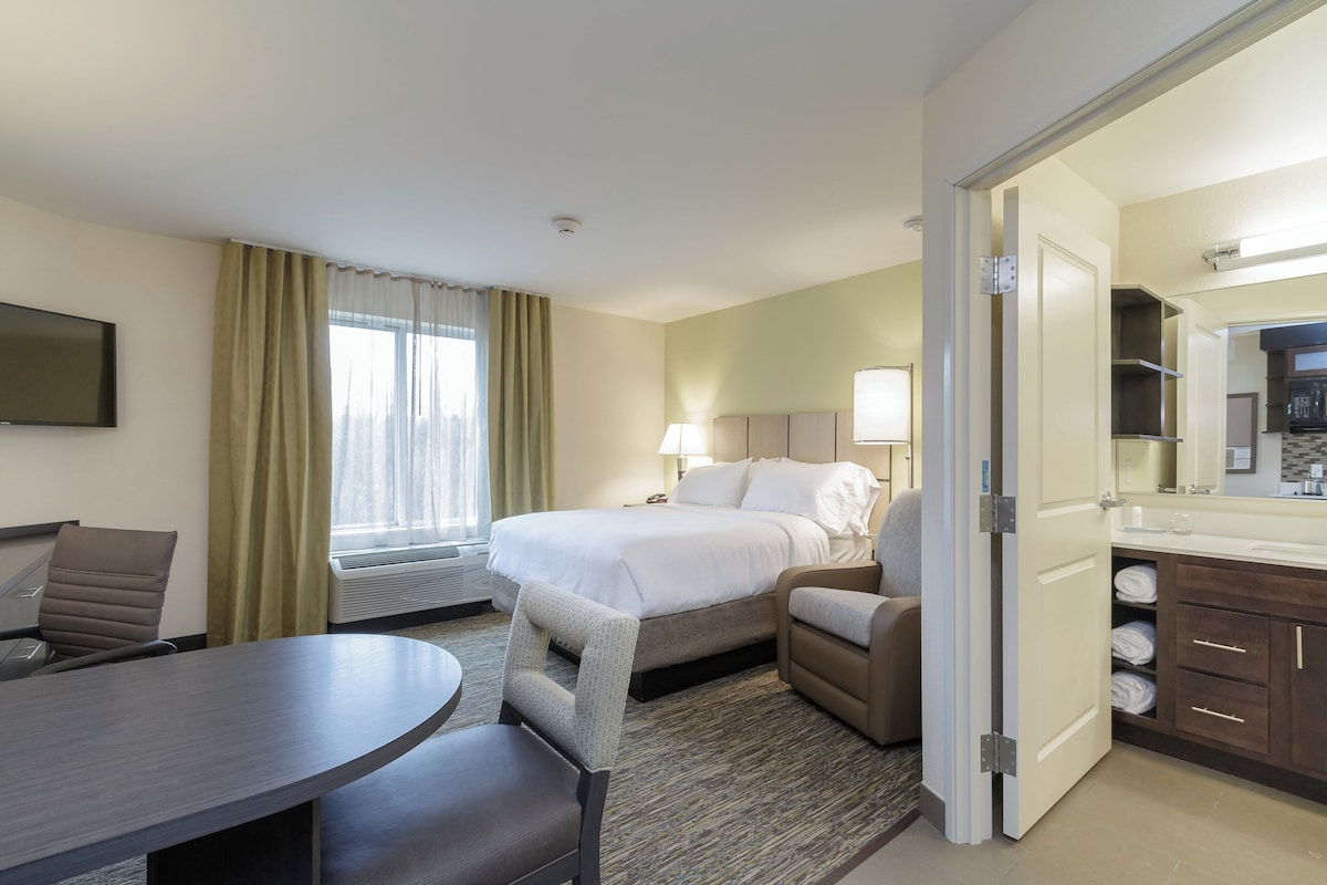 Habitación de hotel con cama, escritorio, silla, mesa de comedor, baño con lavamanos y espejo, y ventana con cortinas.