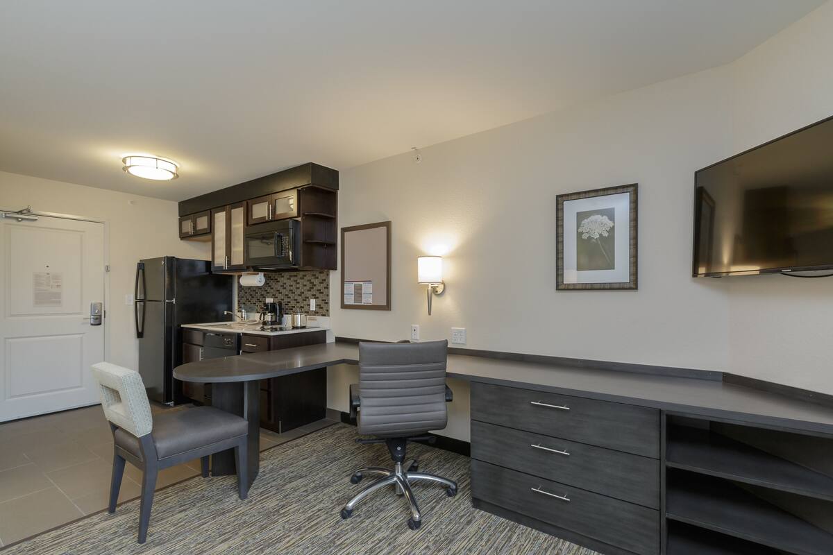 A compact kitchen with modern appliances, a dining area with a chair, and a flat-screen TV mounted on the wall.