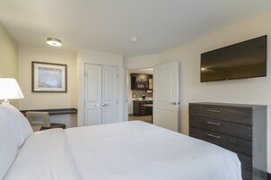 Habitación de hotel con cama, cómoda, televisión y una cocina visible a través de una puerta abierta.