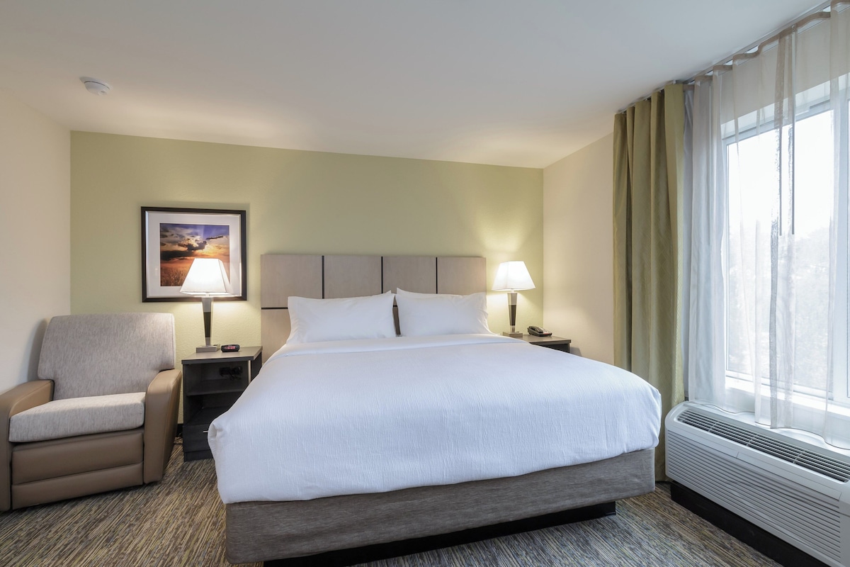 Una habitación de hotel con cama, una silla, una mesita de noche, una lámpara, un cuadro en la pared y una ventana con cortinas.