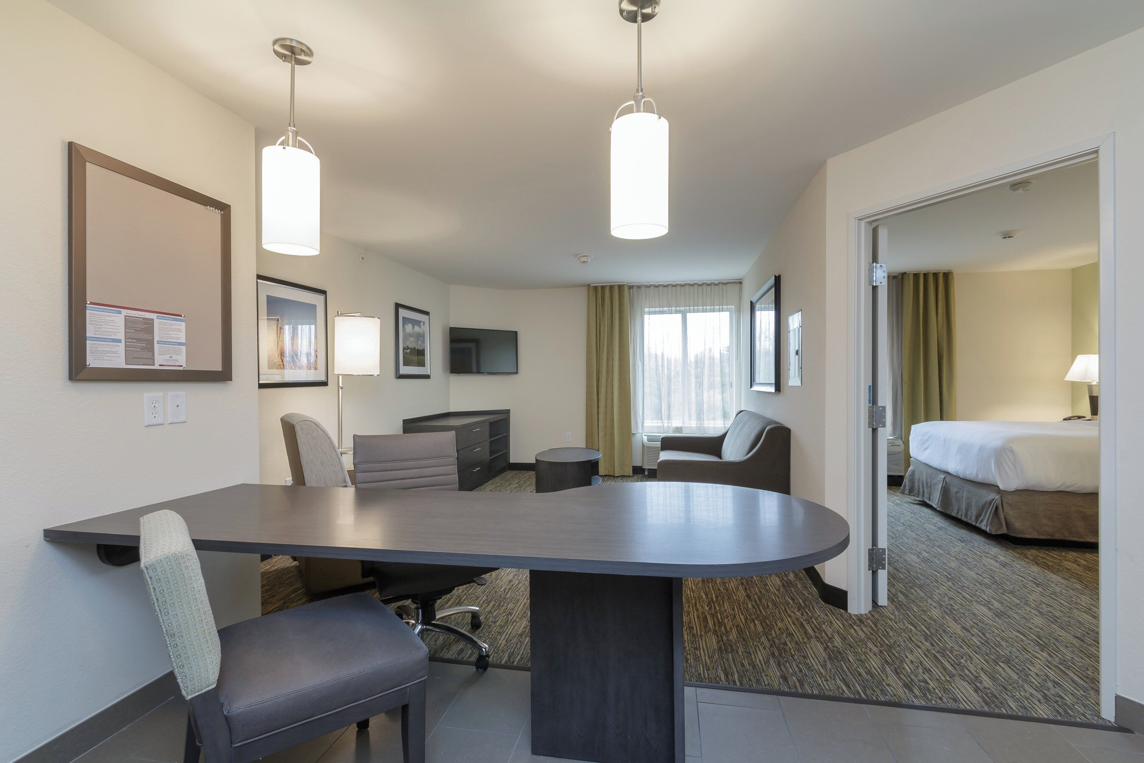 A hotel room with a large desk, two chairs, a bed, a TV, and a view of the outdoors.