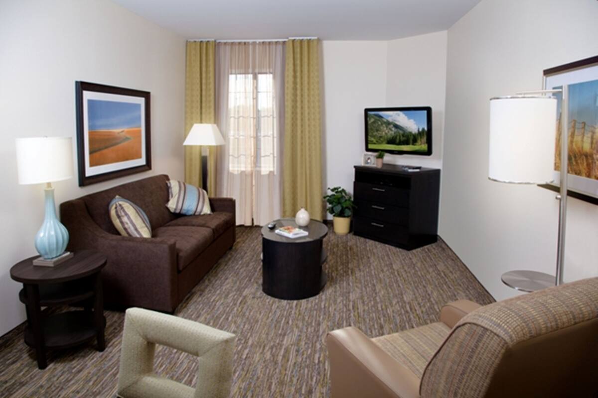 Quarto de hotel com um sofá marrom, uma mesa redonda pequena, uma televisão, um abajur e um quadro na parede.