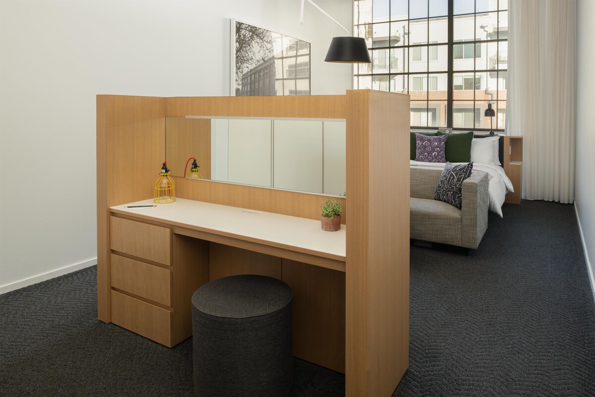 A modern room with a wooden desk, a small stool, a mirror, and a bed with a green cushion.
