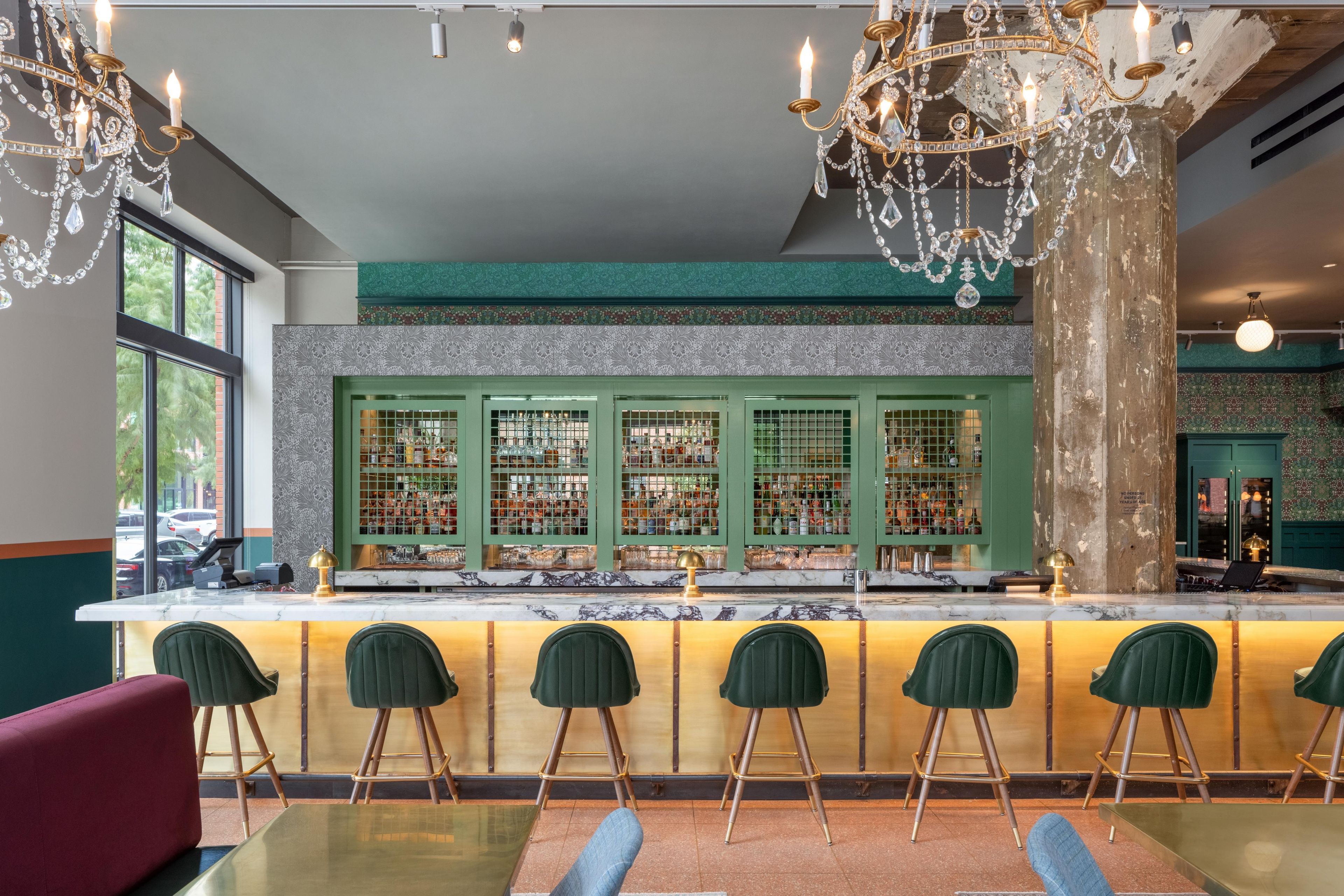 A bar area with a marble countertop, green bar stools, and a decorative chandelier.