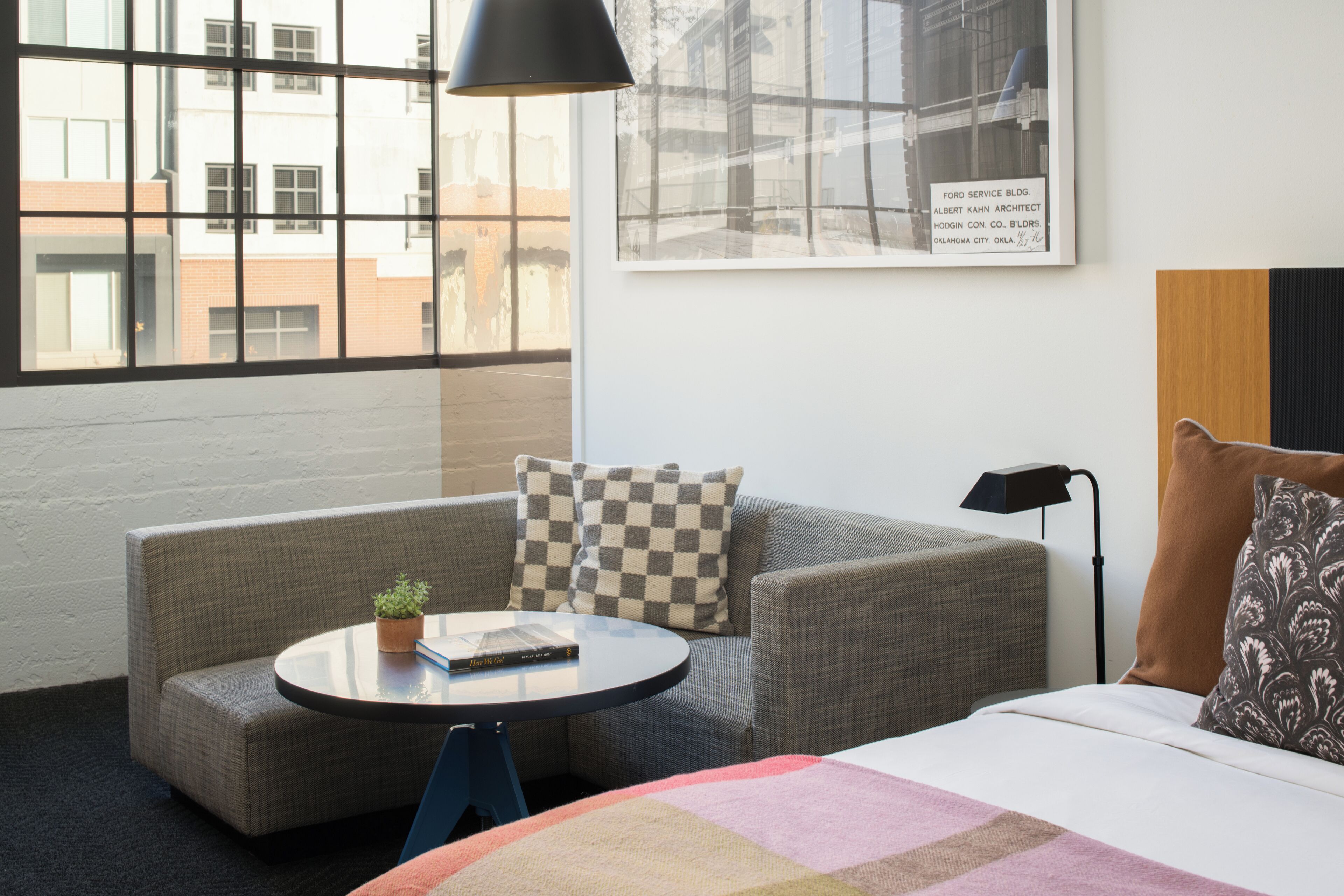 Una habitación de hotel moderna con un ventanal grande, un sofá, una mesita redonda y una cama con una colcha a cuadros.