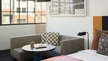 Una habitación de hotel moderna con un ventanal grande, un sofá, una mesita redonda y una cama con una colcha a cuadros.