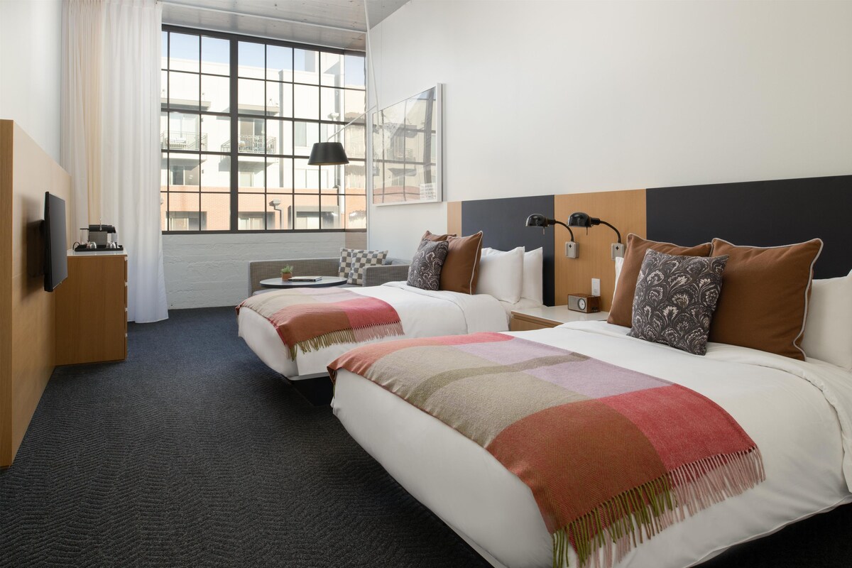 A hotel room with two beds, a desk, a chair, a TV, and a window with a view of buildings.