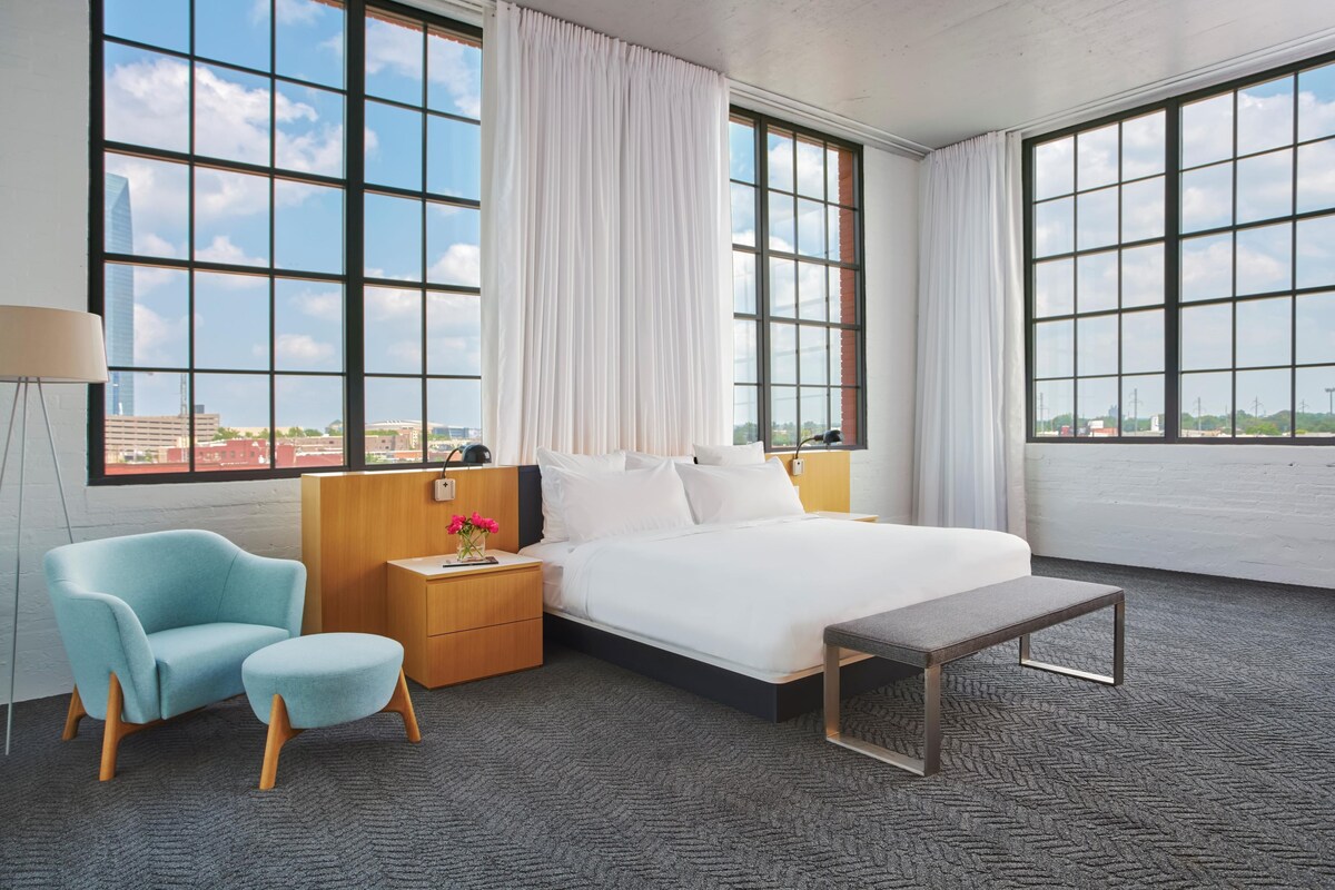 A hotel room with a large bed, a bedside table, a lamp, a chair, and a view of the city through large windows.