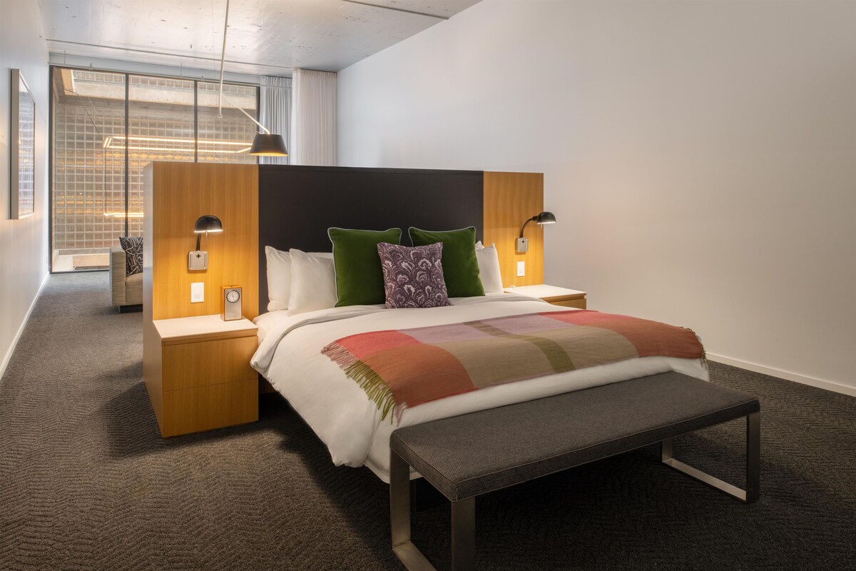 A modern hotel room with a large bed, a bench, a nightstand, a lamp, and a view of a cityscape through the window.