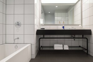 A modern bathroom with a white tiled wall, a black metal bathtub, a black sink with a mirror above, and a towel rack with folded towels.