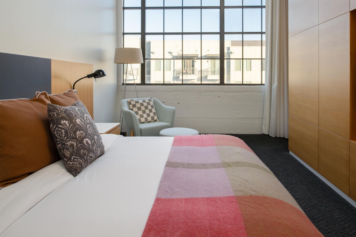 A hotel room with a bed, a chair, a small table, and a window with a view of buildings.