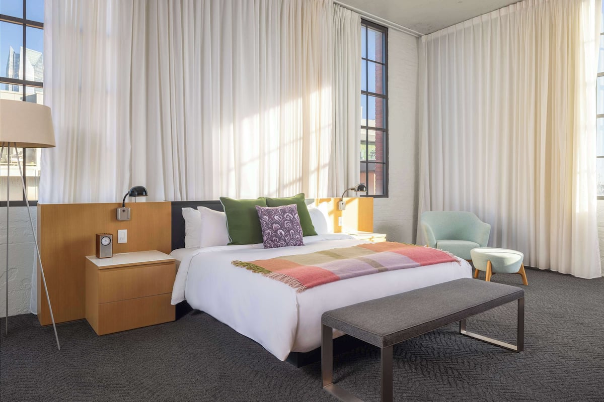 Habitación de hotel con cama, mesitas de noche, un banco, una silla y ventanas grandes con cortinas.
