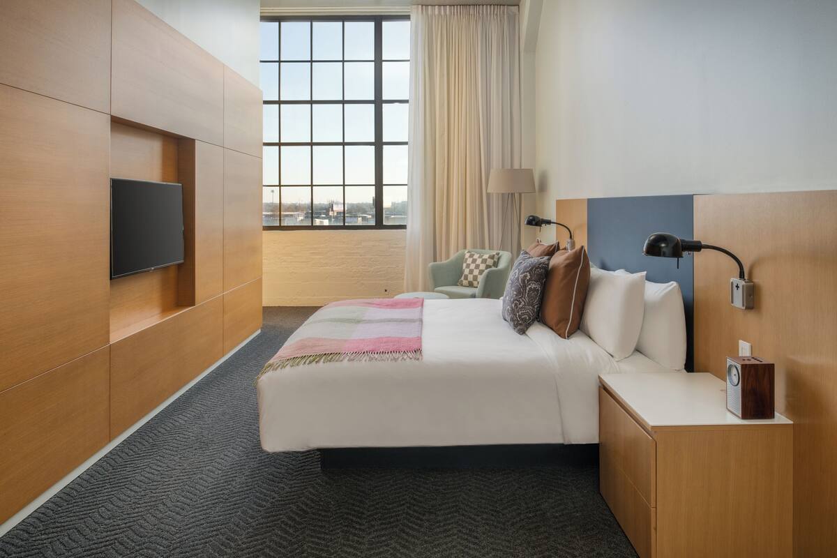 Una habitación de hotel con una cama grande, un televisor de pantalla plana montado en la pared, una mesita de noche con lámpara y radio, y vistas a la ciudad a través de amplias ventanas.