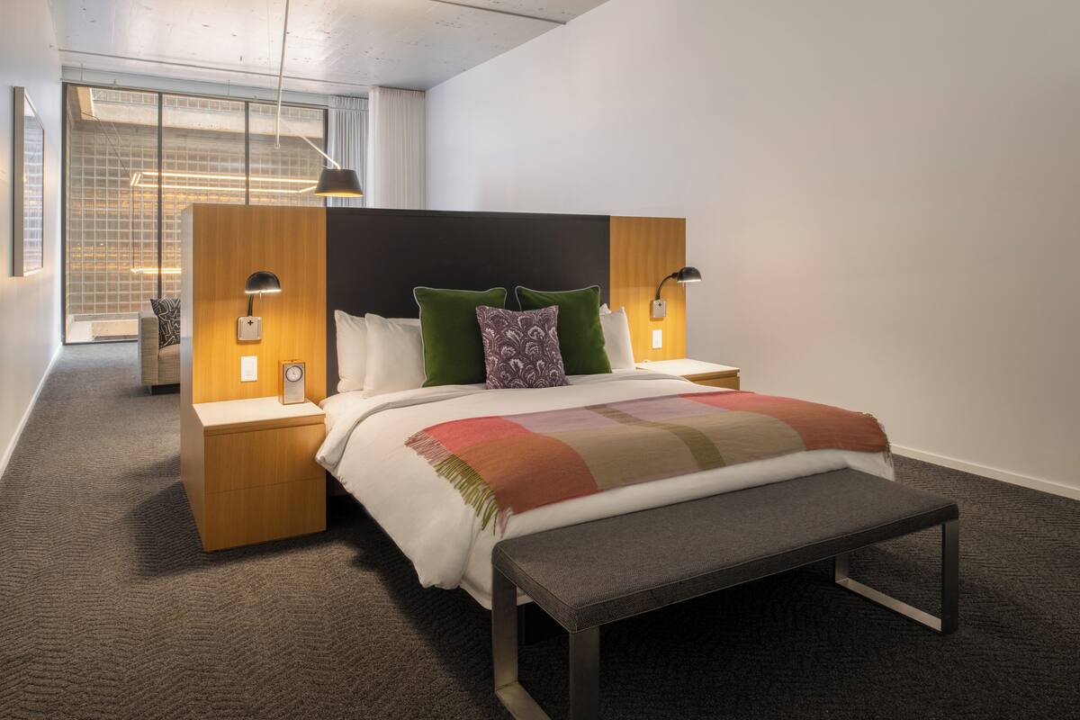 Habitación de hotel moderna con una cama grande, dos mesitas de noche, un banco y vistas a la ciudad a través de amplios ventanales.