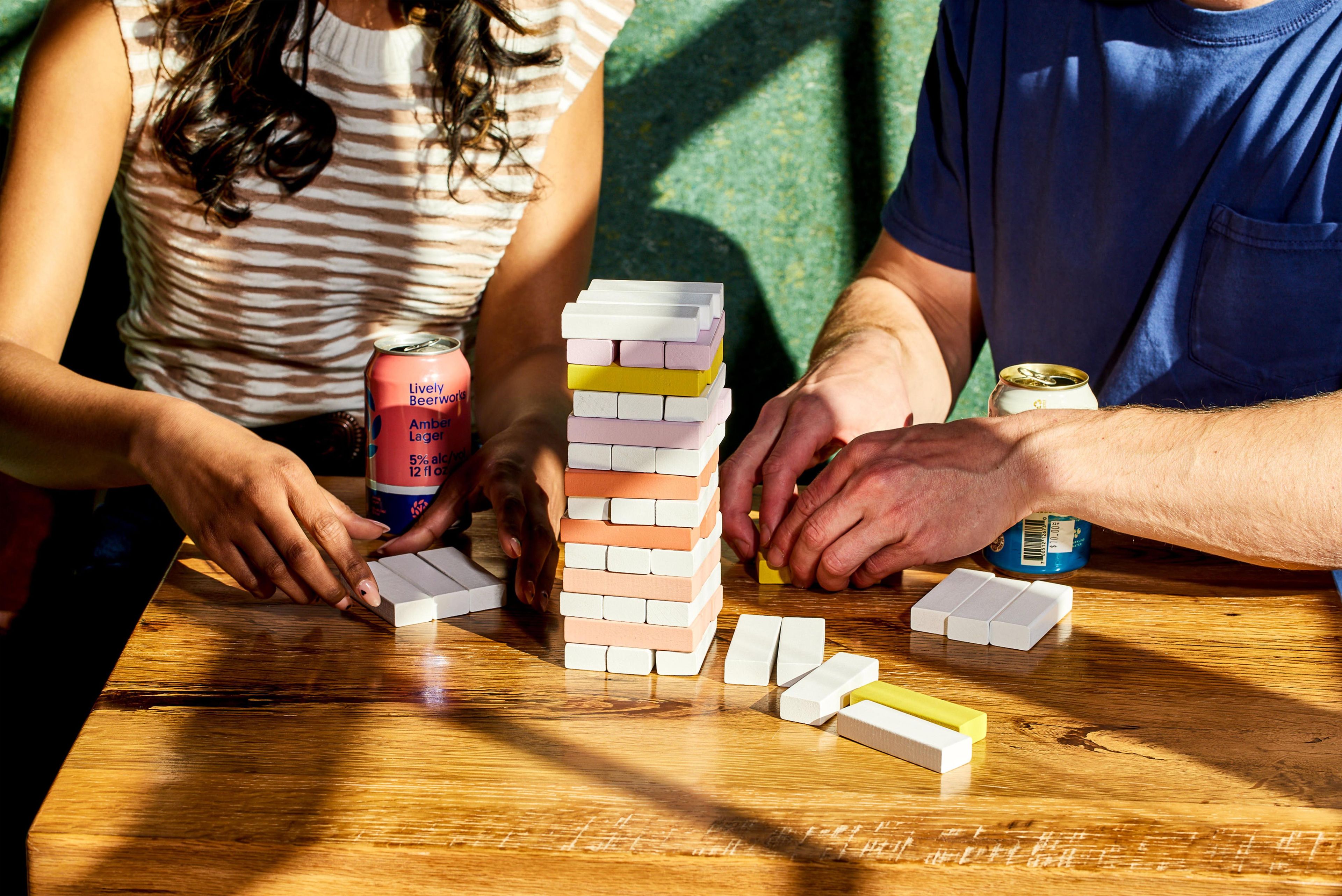 Two people playing a game with stacked blocks and cans of beer on a wooden table.