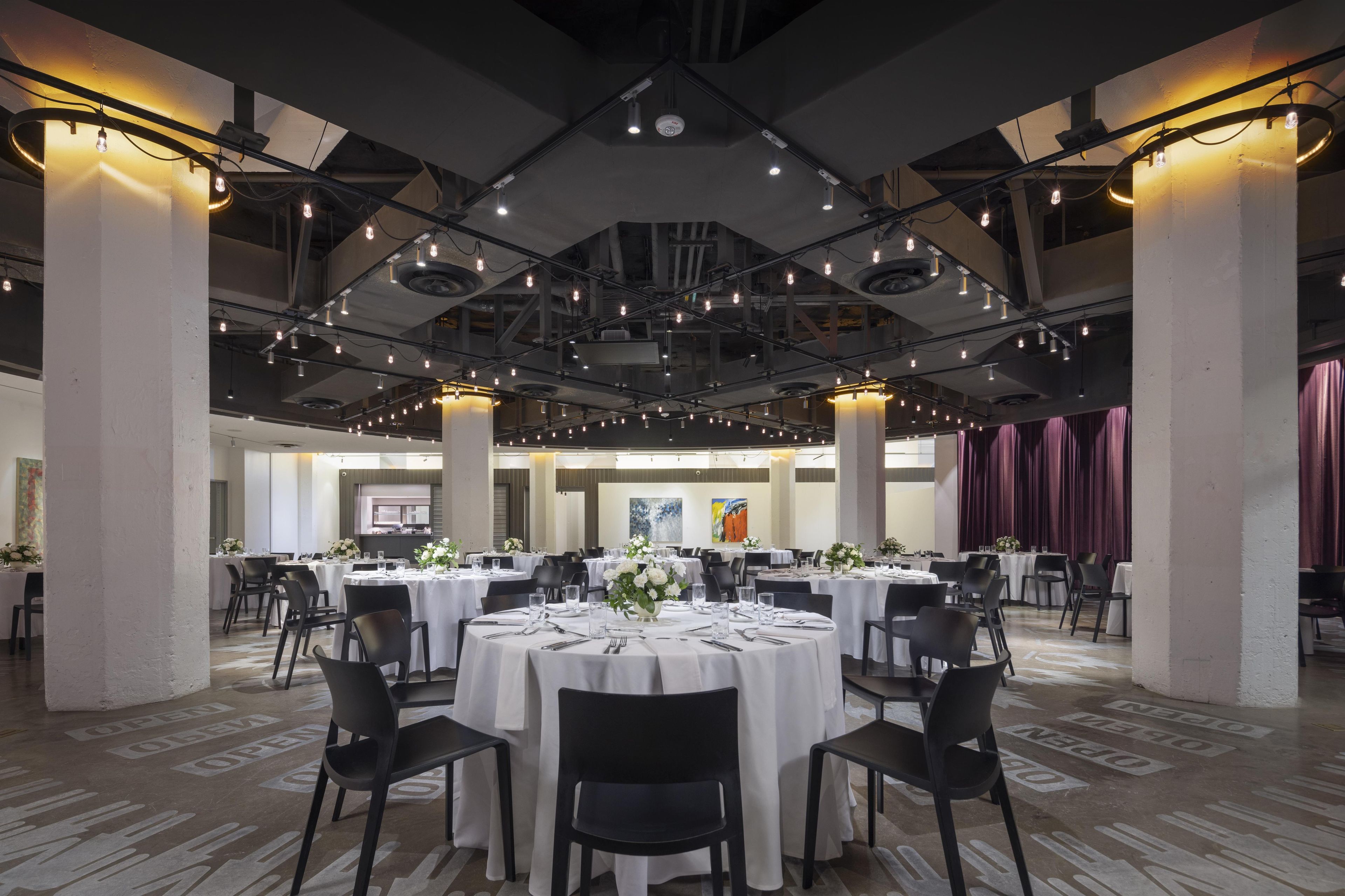Un amplio salón de banquetes con mesas redondas dispuestas para comer, techos altos con vigas a la vista e iluminación moderna.