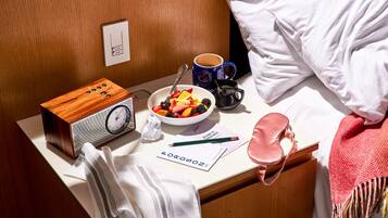 A neatly arranged hotel room with a wooden bedside table, a radio, a bowl of fruit, a coffee mug, a tissue, a welcome brochure, a pink bag, and a white pillow.