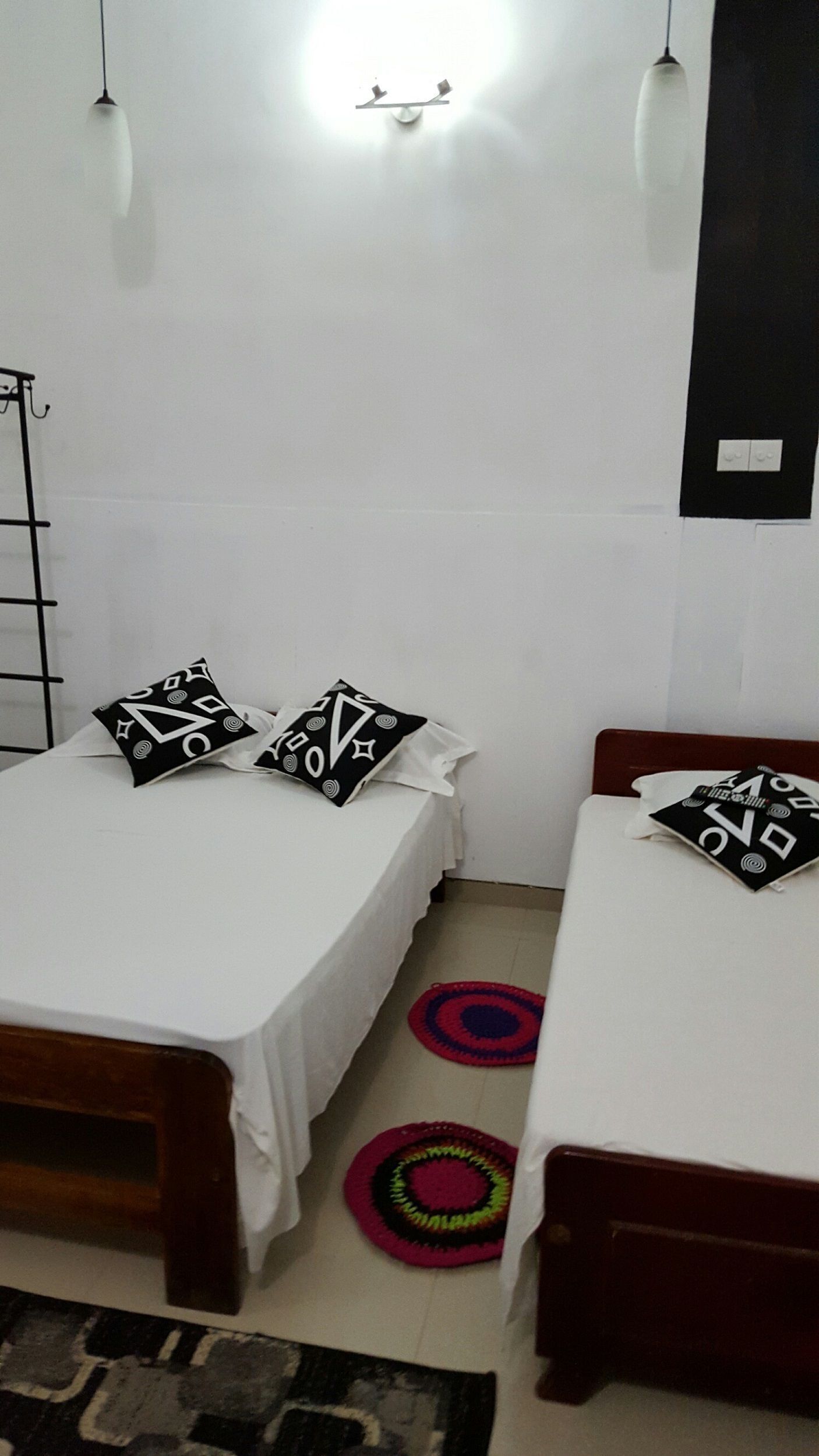 A bedroom with two beds, each with a wooden frame and white bedding. There are two black and white patterned pillows on each bed. The room has a white wall with a black switchboard and a white ceiling light fixture.