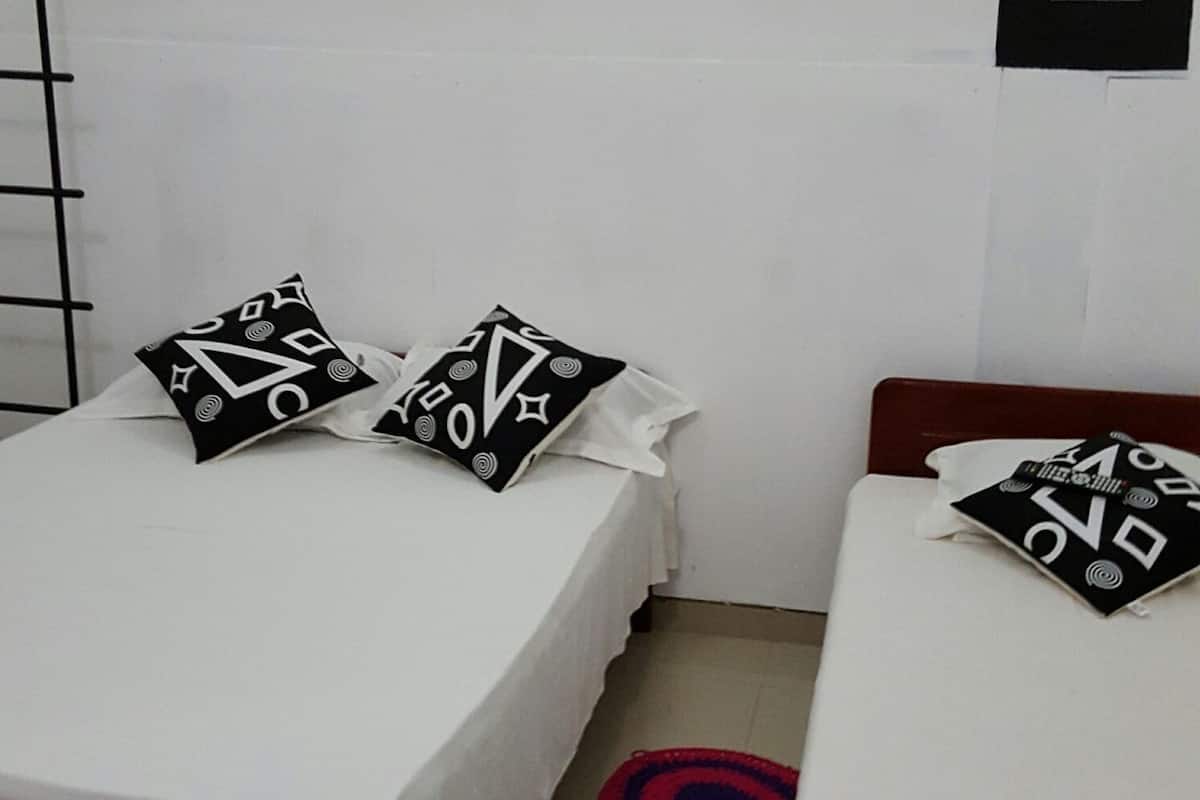A bedroom with two beds, each with a wooden frame and white bedding. There are two black and white patterned pillows on each bed. The room has a white wall with a black switchboard and a white ceiling light fixture.