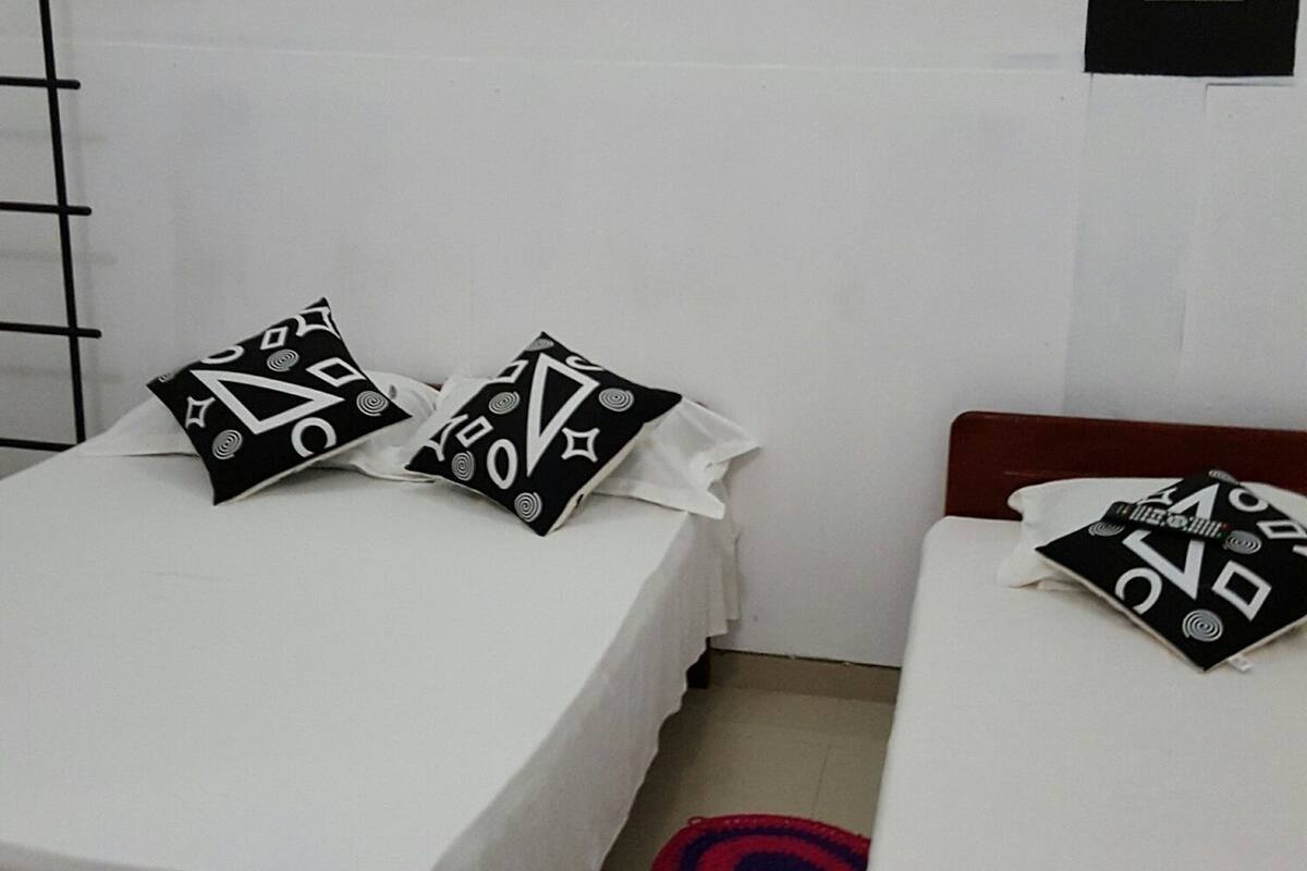 Un dormitorio con dos camas, cada una con estructura de madera y ropa de cama blanca. Hay dos almohadas estampadas en blanco y negro sobre cada cama. La habitación tiene una pared blanca con un panel de interruptores negros y un accesorio de luz blanca en el techo.