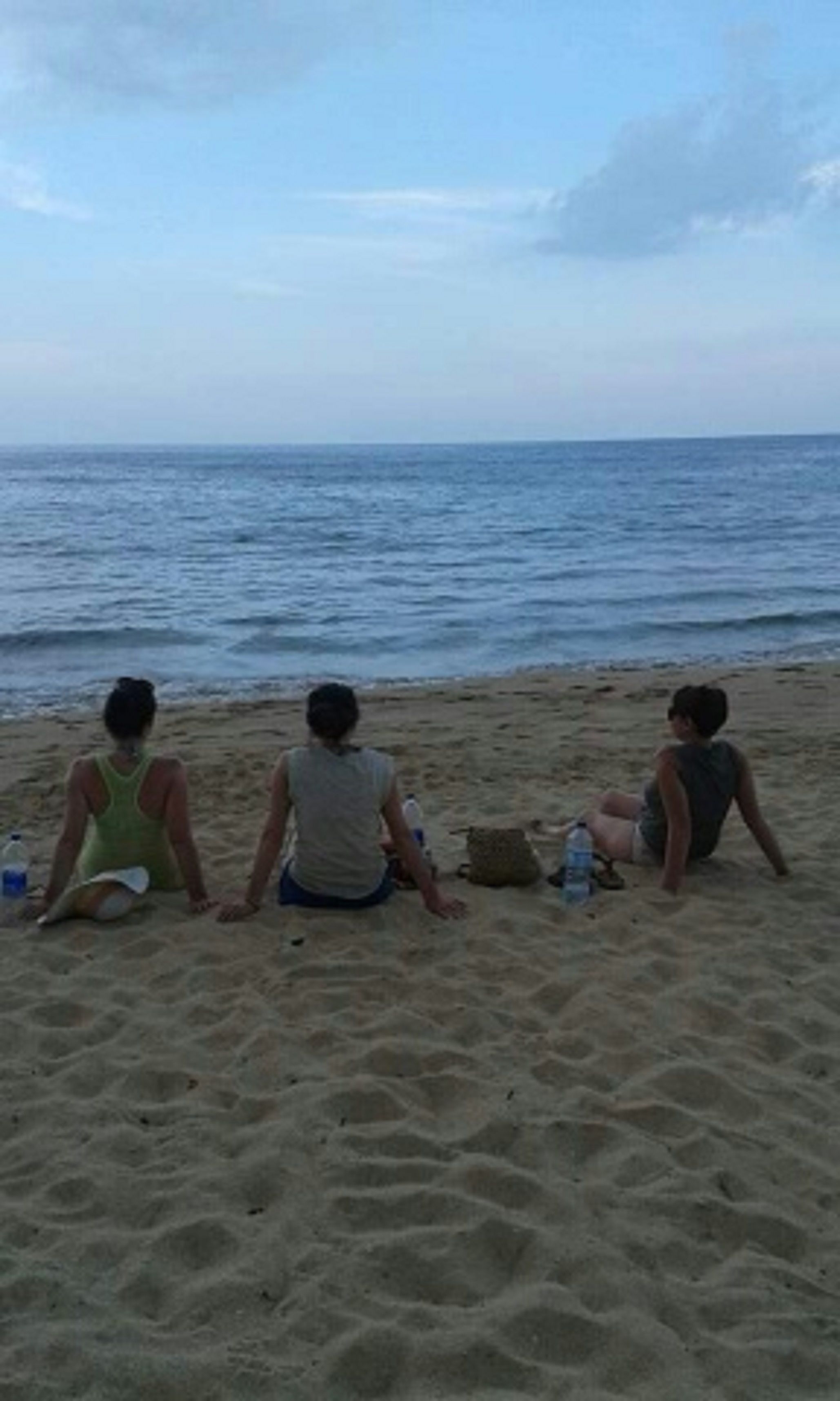 Three individuals sitting on a sandy beach facing the ocean.