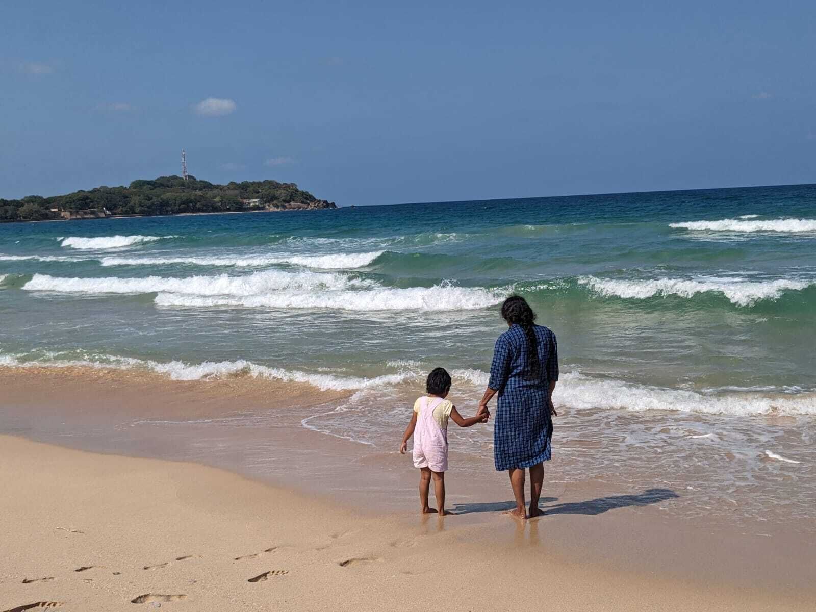 Escena de playa con una persona y un niño tomados de la mano, mirando hacia el océano.