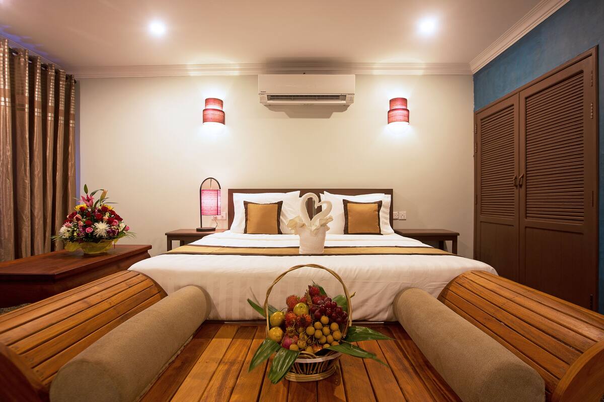 A hotel room with a large bed, two bedside tables, a wooden bench, a fruit basket, and a wall-mounted air conditioner.