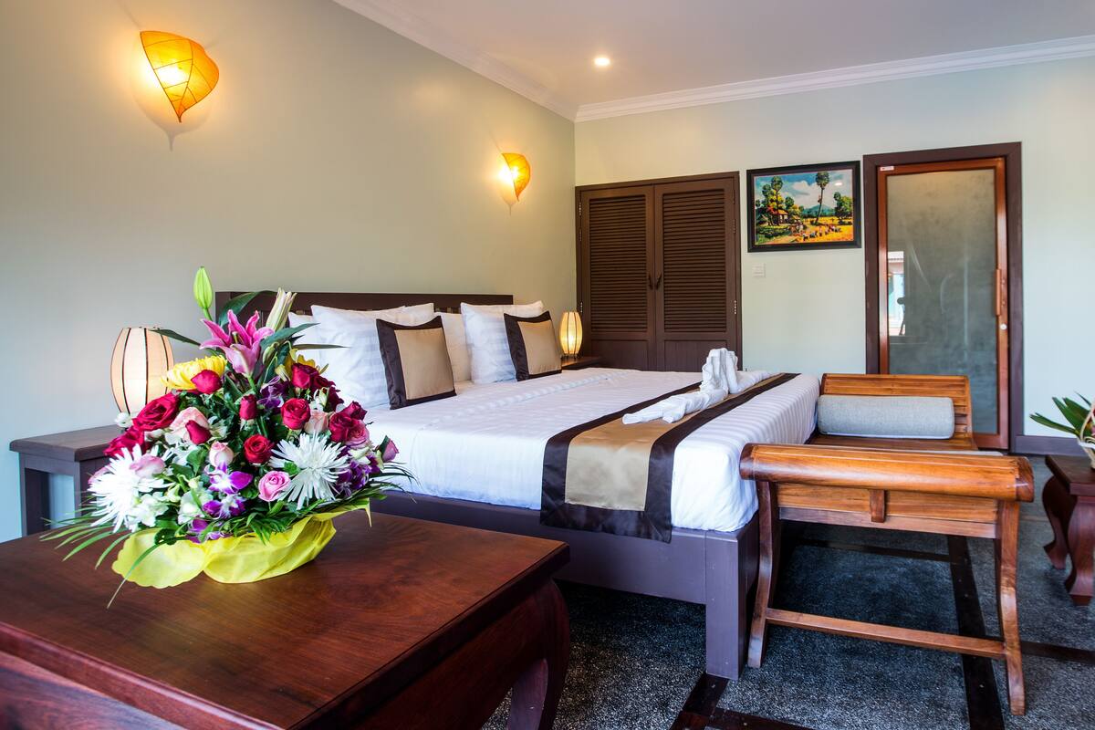 A hotel room with a bed, bedside tables, a desk, a chair, a wooden bench, a fruit basket, and a painting on the wall.