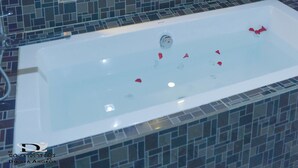 Une salle de bain avec un grand miroir, une baignoire remplie de pétales de roses, une chaise et un vase avec des plantes.