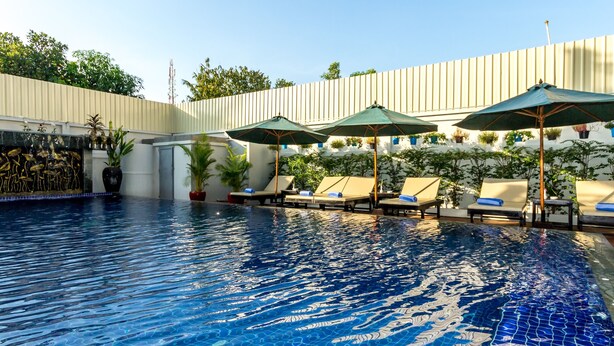 Une piscine entourée d’un mur blanc et de verdure, avec des chaises longues et des parasols.