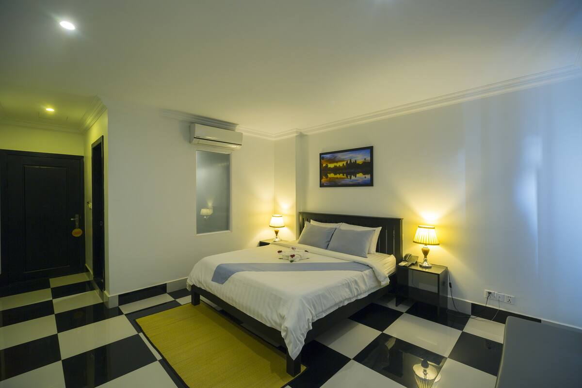 A hotel room with a checkered floor, a bed with white linens, two bedside lamps, a painting on the wall, and an air conditioning unit.