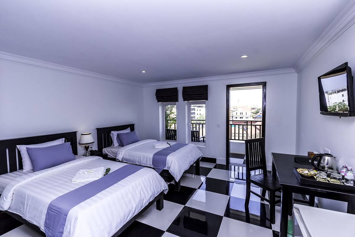 A hotel room with two beds, a dining table, a chair, and a balcony with a view of buildings.