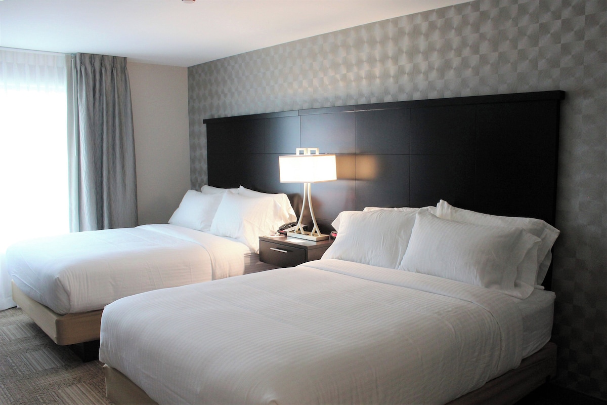 A hotel room with two beds, a nightstand with a lamp, and a large headboard.