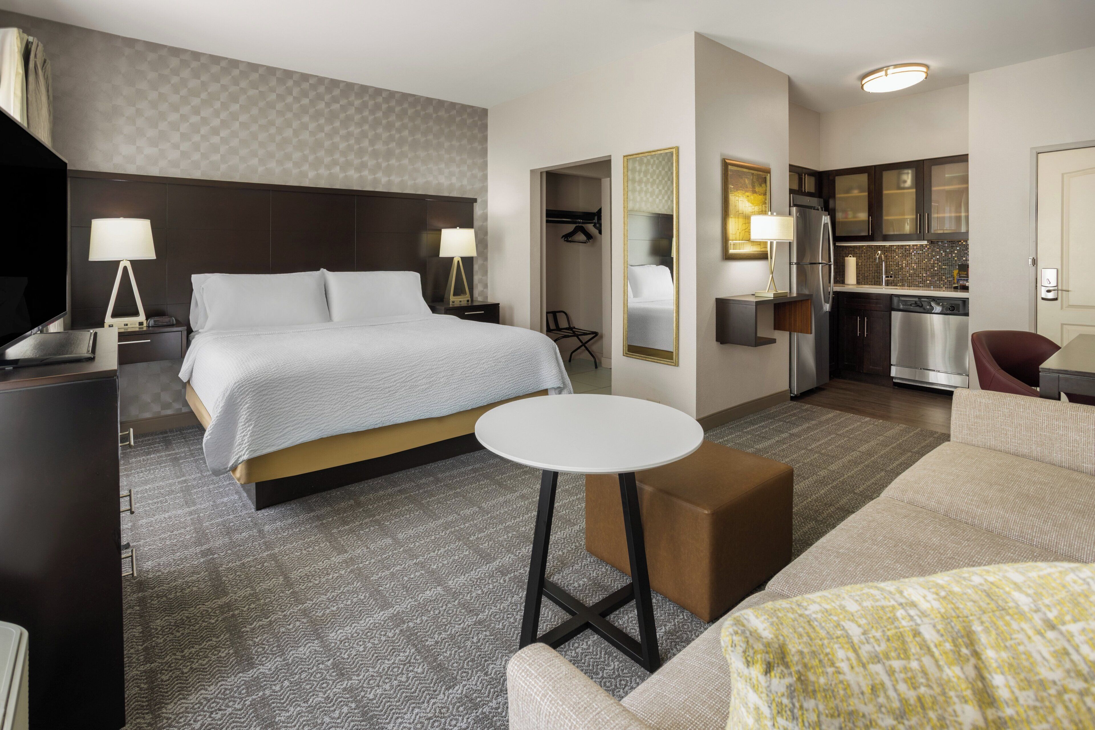 A hotel room with a bed, bedside tables, a small round table, a sofa, a kitchenette, and a TV.