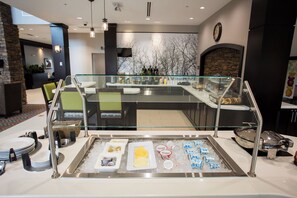 A buffet area with a glass barrier, various food items, and a clock on the wall. - Staybridge Suites Miamisburg by IHG (Miamisburg)
