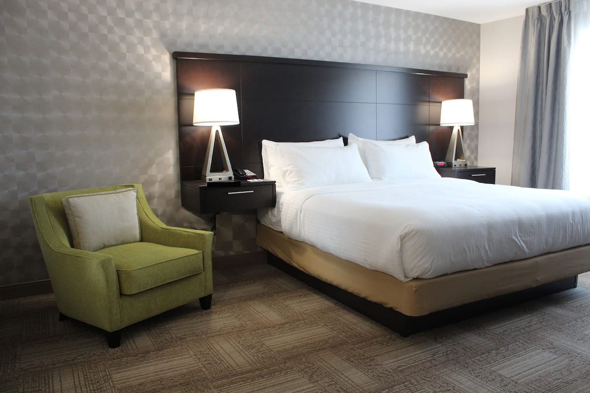 A hotel room with a large bed, two bedside tables with lamps, a green armchair, and a patterned wall.
