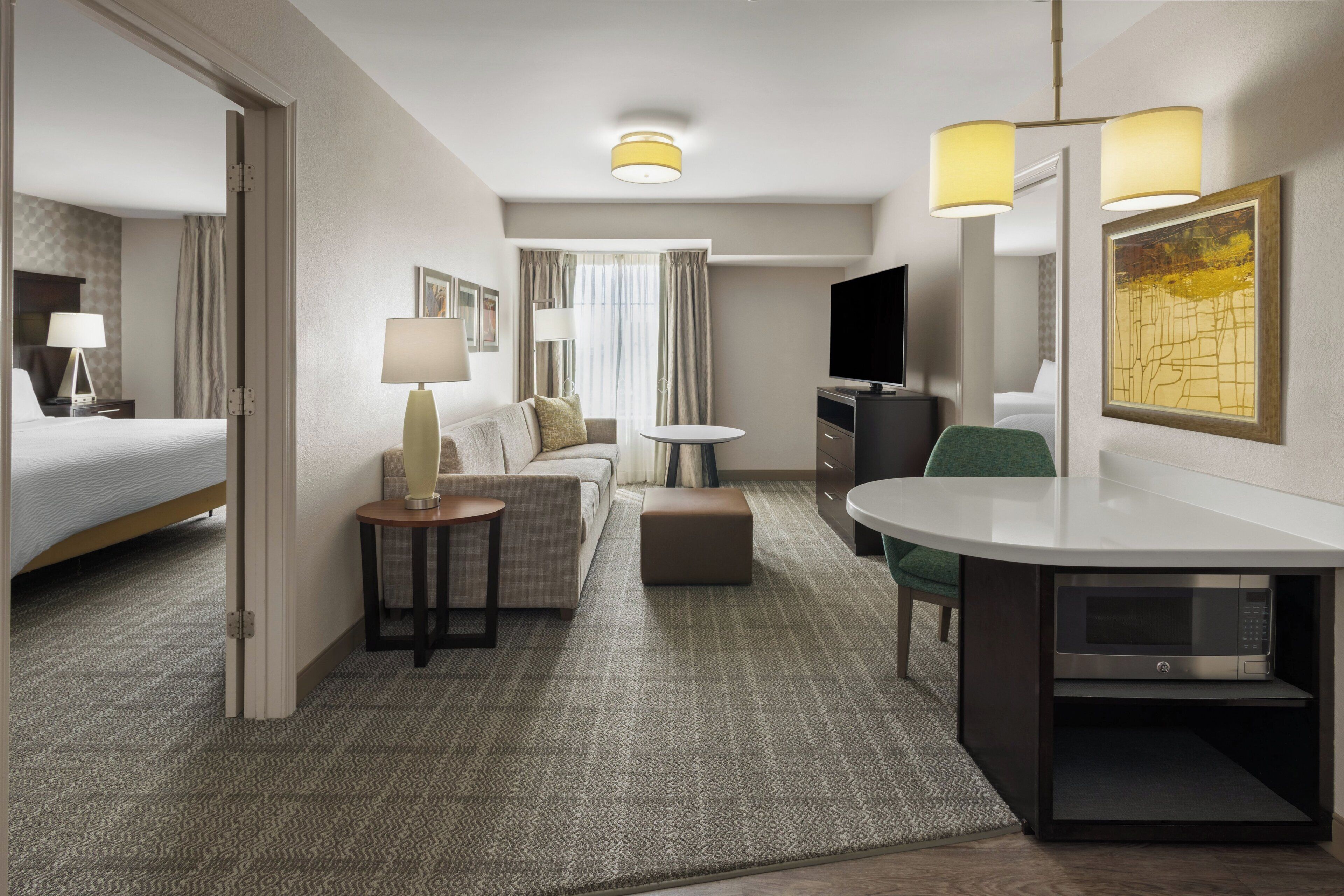 A hotel room with a bed, a sofa, a desk with a chair, a TV, and a painting on the wall.