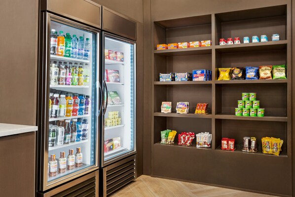 A refrigerated beverage cooler stocked with various drinks and a shelf unit with snacks and packaged goods. - Staybridge Suites Miamisburg by IHG (Miamisburg)