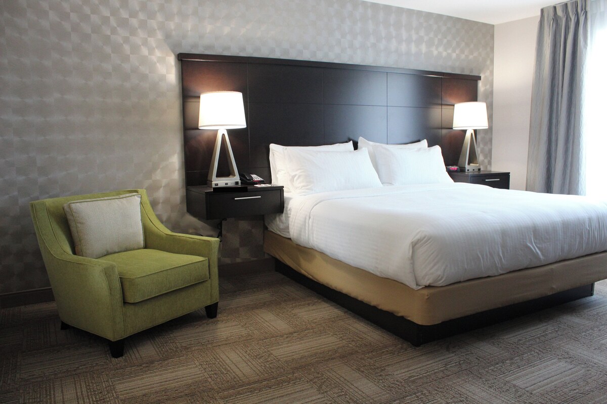 A hotel room with a large bed, two bedside tables with lamps, a green armchair, and a patterned wall.