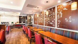 Un restaurante con mesas de madera y sillas rojas, una zona de bar y una pared decorada con ilustraciones culturales.