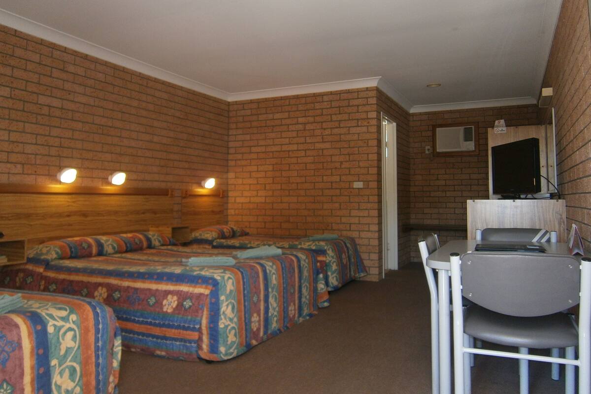 A room with brick walls, a wooden headboard, a desk with a chair, and a television on the wall.
