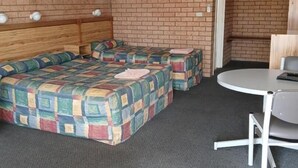 A room with a brick wall, a wooden headboard bed, a colorful quilted bedspread, a small table with a chair, and an air conditioning unit. - Aaron Inn Motel (Narrabri)