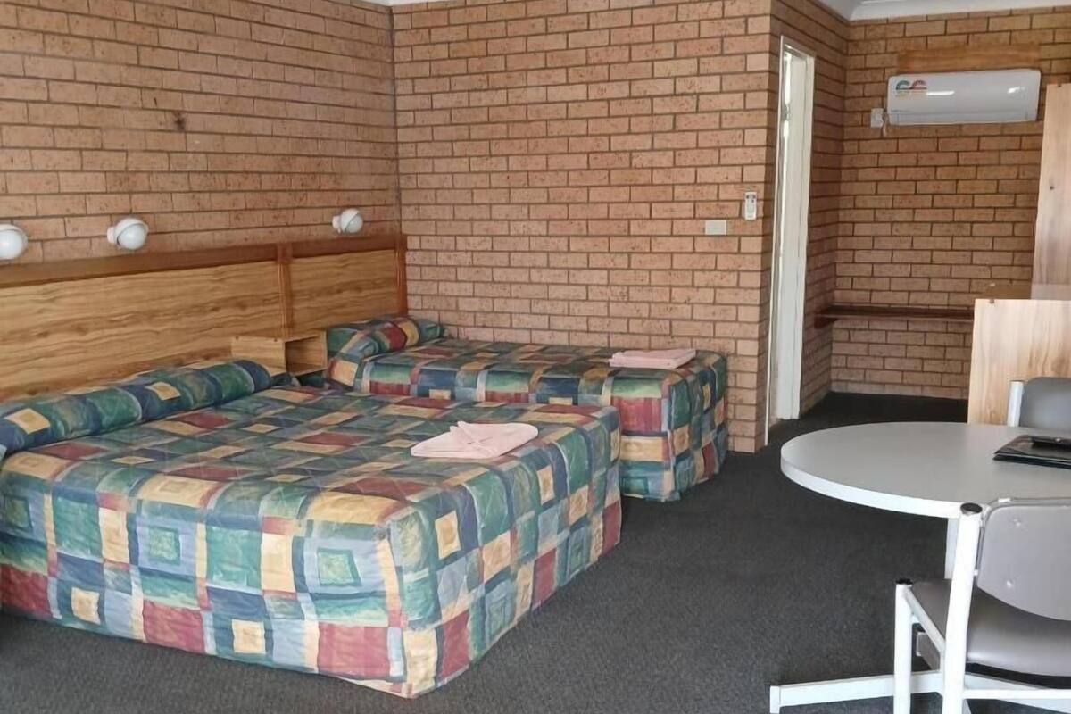 A room with a brick wall, a wooden headboard bed, a colorful quilted bedspread, a small table with a chair, and an air conditioning unit.