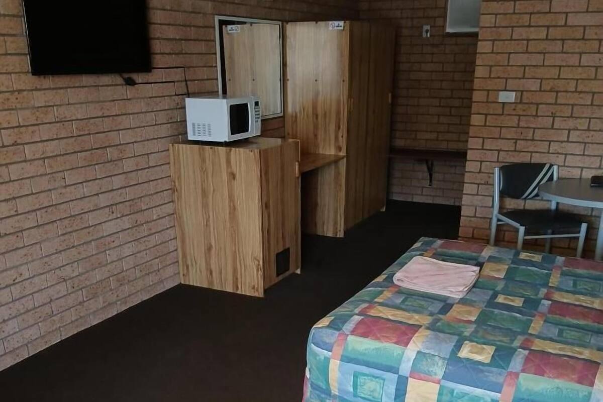 A hotel room with a brick wall, a bed with a colorful quilt, a wooden wardrobe, a small table, and a television mounted on the wall.