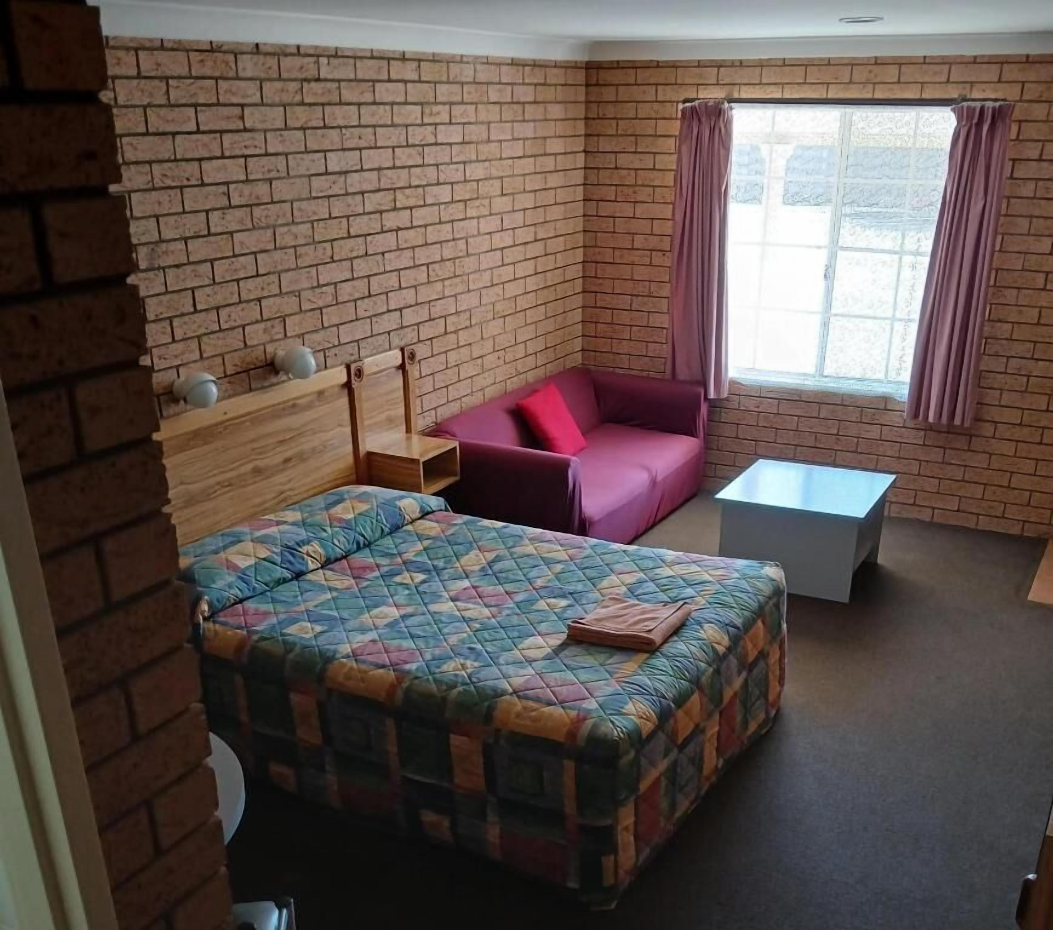 A room with a brick wall, a bed with a colorful quilt, a red sofa, a small table, and a window with curtains.