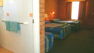 Une chambre d’hôtel avec une salle de bain carrelée de carreaux blancs, un porte-serviettes et un mur en briques. La chambre comprend trois lits avec une literie bleue et une fenêtre avec des rideaux.