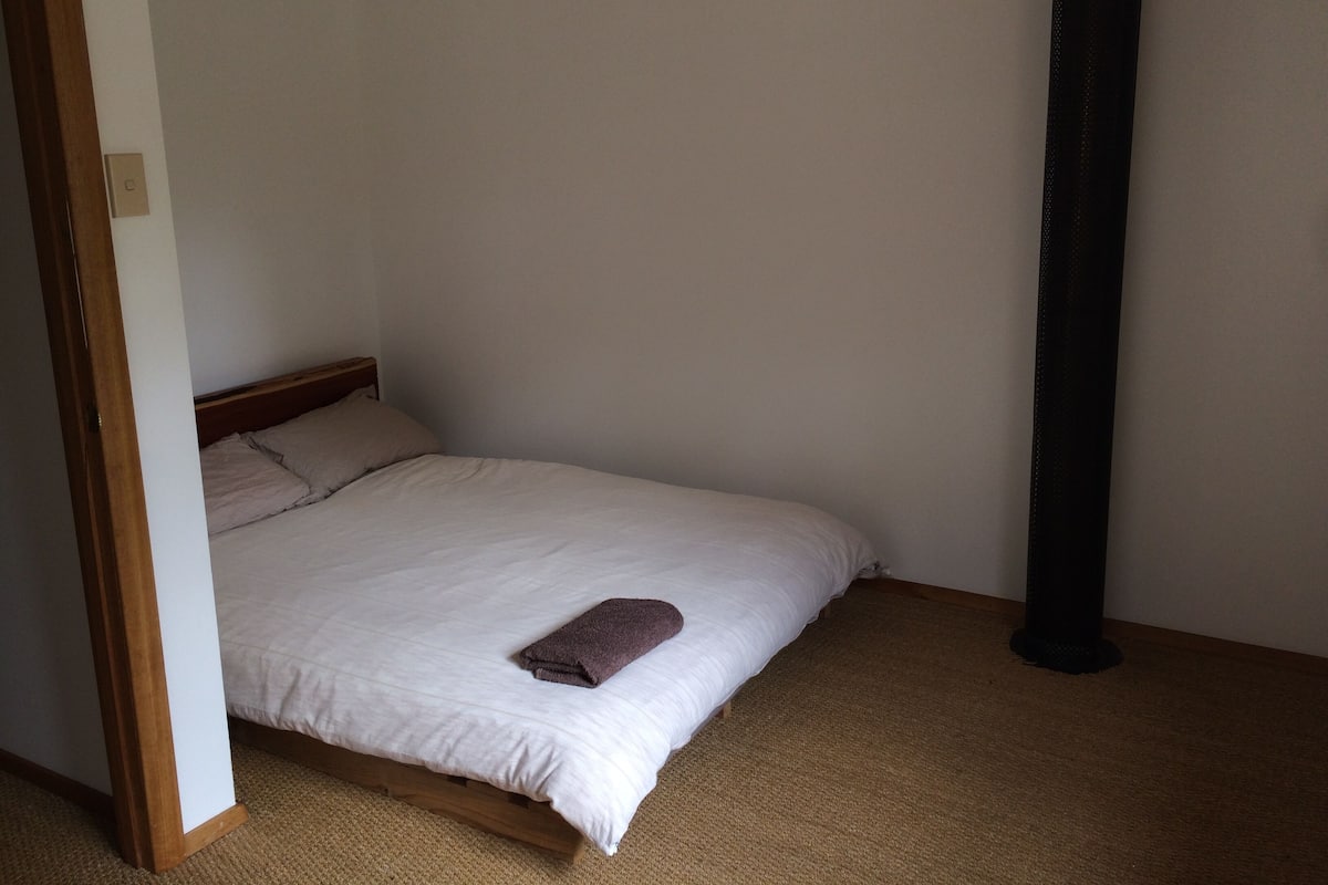 A single bed with a white mattress and two pillows, a brown towel on the bed, and a tall black object to the right.