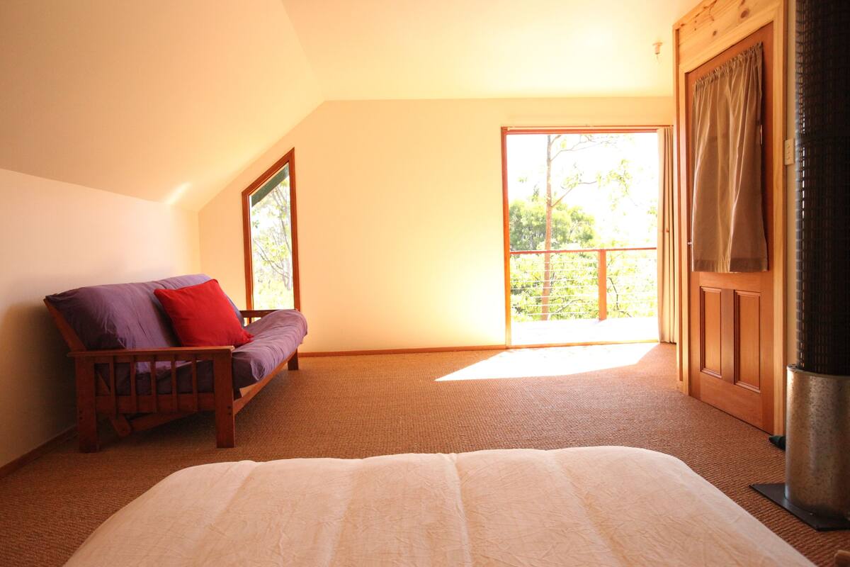 A bedroom with a bed, a wooden chair with cushions, a large window, and a door leading outside.