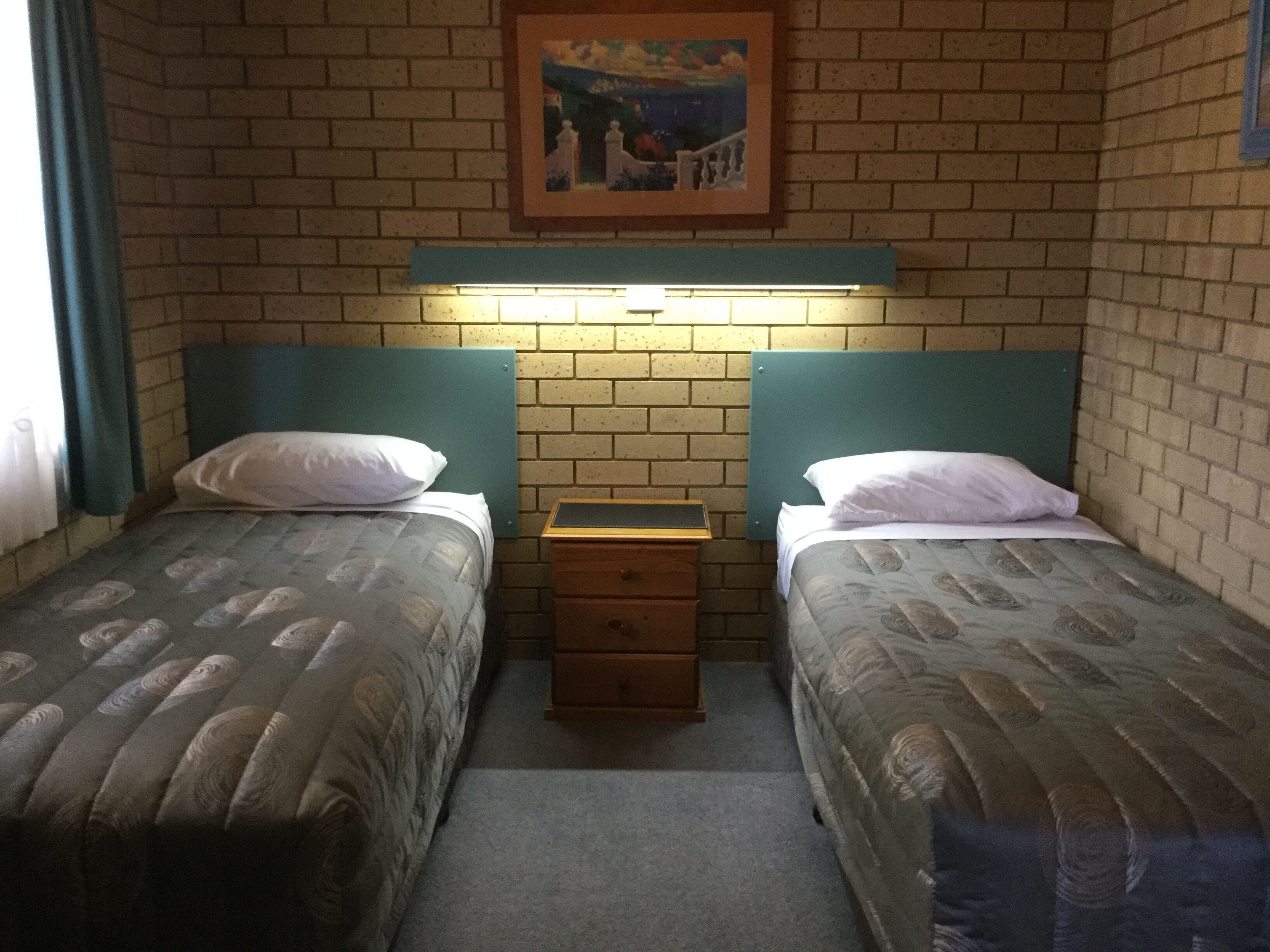 Quarto com duas camas de solteiro, mesa de cabeceira de madeira, quadro pendurado na parede e parede de tijolos.