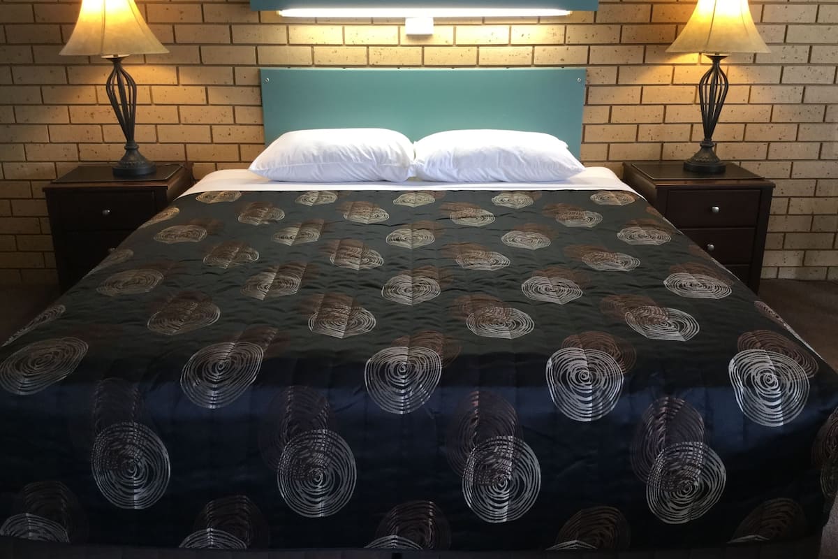 A neatly made bed with a patterned bedspread, two bedside lamps, and a headboard against a brick wall.